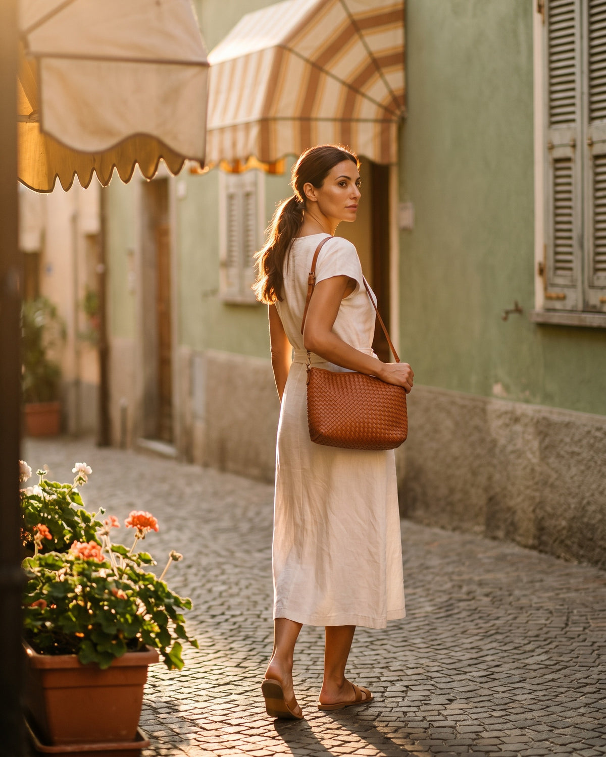 Donna che cammina in un vicolo al tramonto con una borsa intrecciata color cognac a tracolla