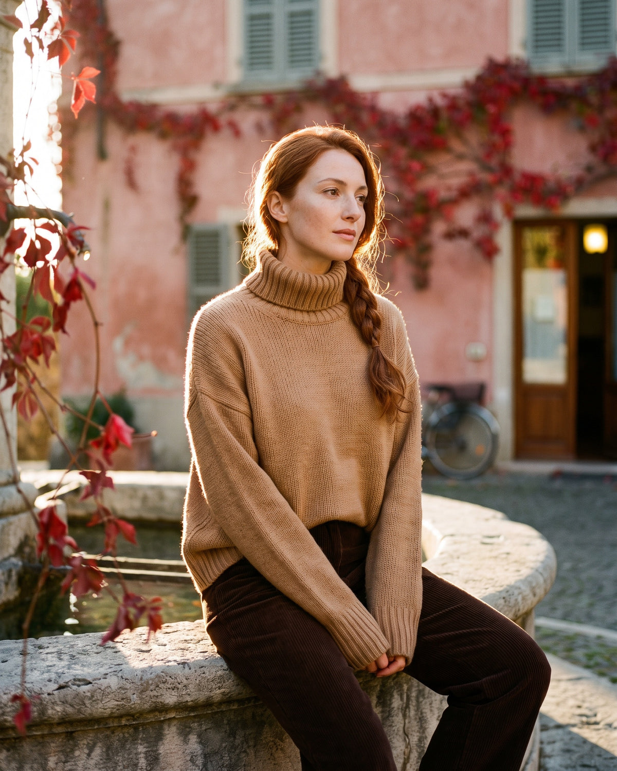 Donna con maglione oversize color cammello seduta sul bordo di una fontana in una piazza italiana al tramonto