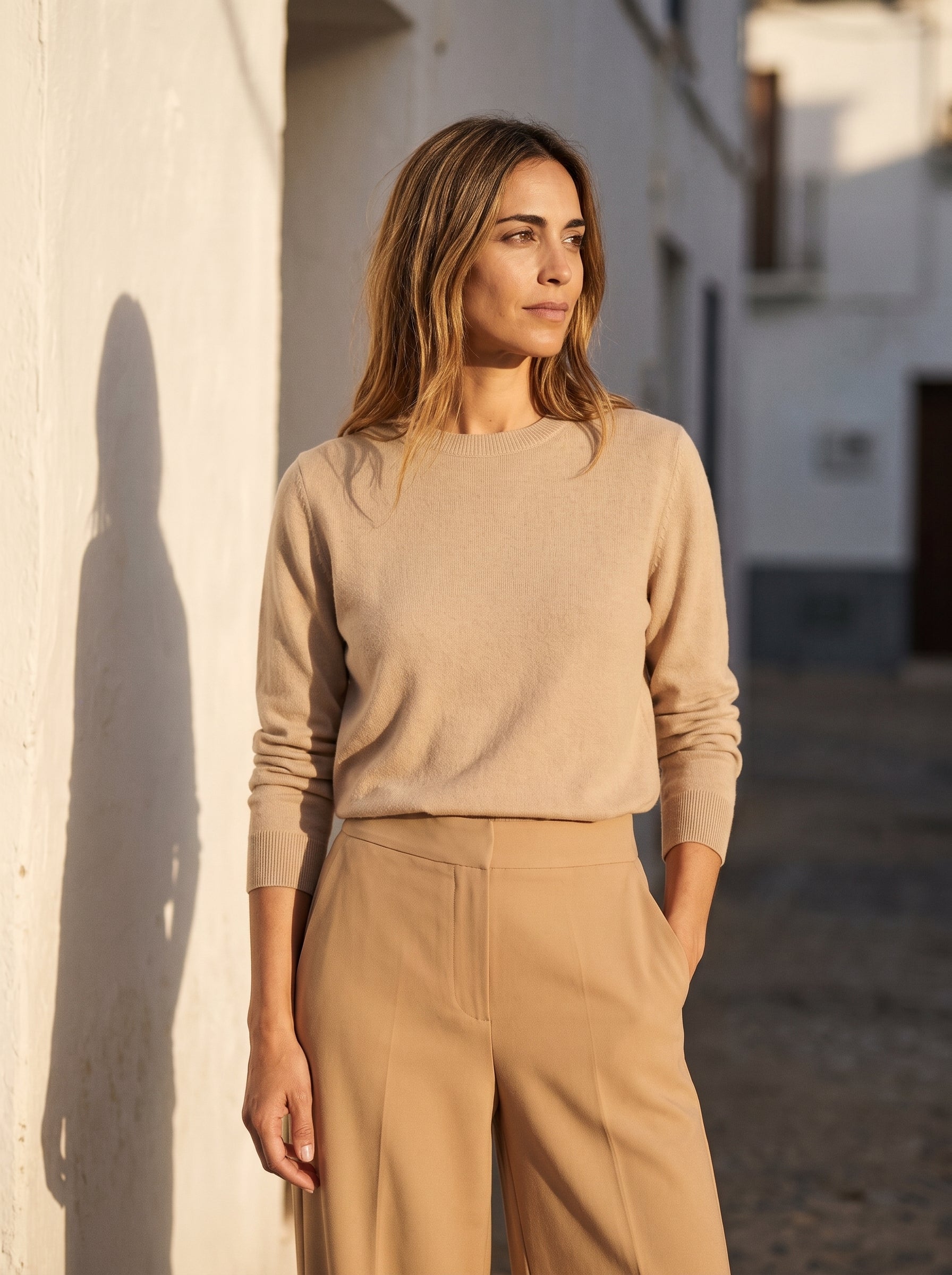 Mujer con jersey beige y pantalón camel en una calle encalada, a última hora de la tarde